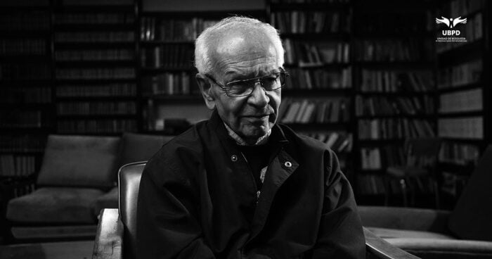 Black-and-white portrait of an elderly man with glasses seated in a library, with shelves of books in the background. - Las2orillas.co: Historias, voces y noticias de Colombia