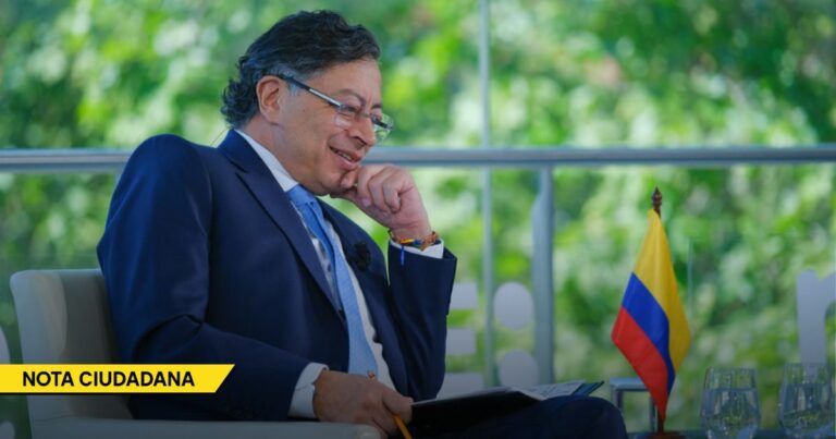 Professional man in a dark blue suit sits smiling and writes in a notebook, with a small Colombian flag on the table and a yellow Nota Ciudadana banner in the corner. - Petro se dedicó a gobernar mirando los errores de sus antecesores, sin responder por los propios