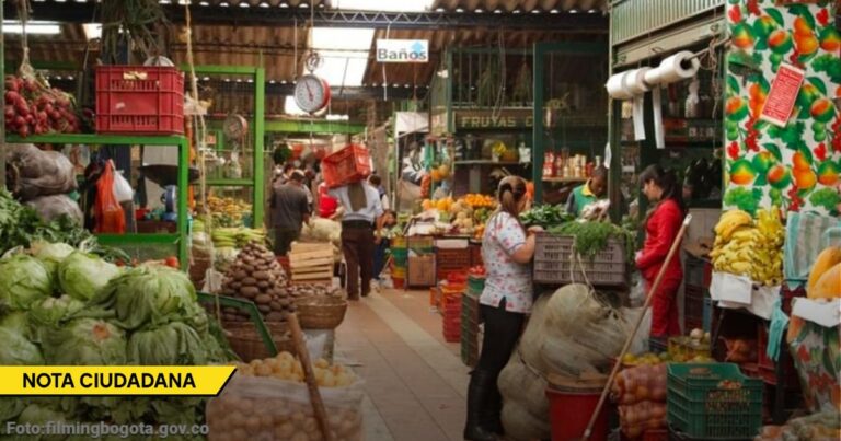Indoor bustling produce market with leafy greens, fruits, and workers behind stalls as customers shop for groceries. - Mientras el gobierno celebra el dólar bajo, la guerra entre Irán e Israel le pega directo al bolsillo de los colombianos