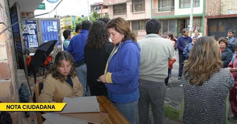 A woman sits at a wooden table signing a notebook as another woman in a blue jacket stands nearby at a street-side civic event, with a crowd and a NOTA CIUDADANA banner in the corner. - Libros perdidos y trasteo de votos, la antesala de la pelea por el botín de las Juntas de Acción Comunal