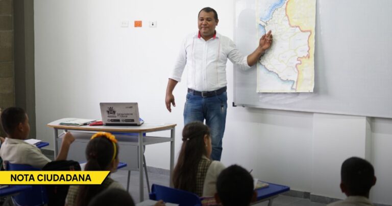 Male teacher presents a large map on a board to students in a classroom, with a laptop on the desk nearby. - El nuevo plan educativo promete mucho pero se estrella con viejos problemas