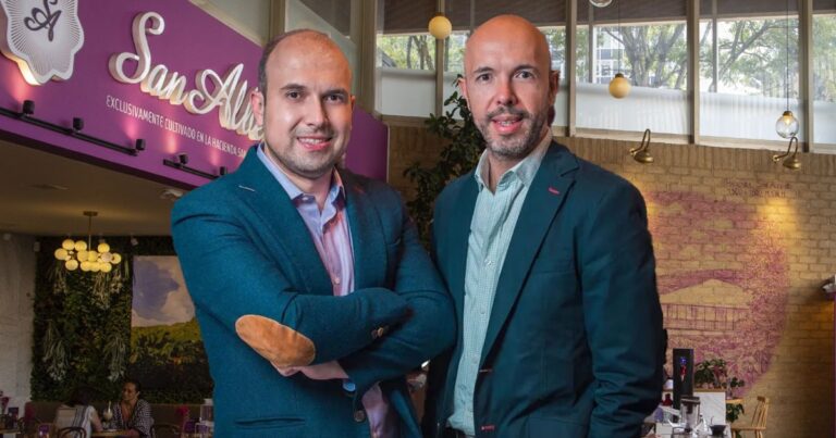 Two smiling men in blue blazers pose in a cafe, arms crossed, with a purple wall and logo in the background. - Un café cultivado en la ladera del departamento del Quindío fue el preferido de los restauranteros colombianos