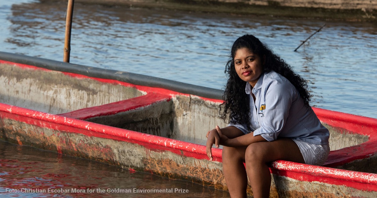 Woman with curly hair sits on a weathered red boat by calm water, looking at the camera. - Una joven ambientalista resultó premiada por defender el suelo de Puerto Wilches de las gigantes petroleras