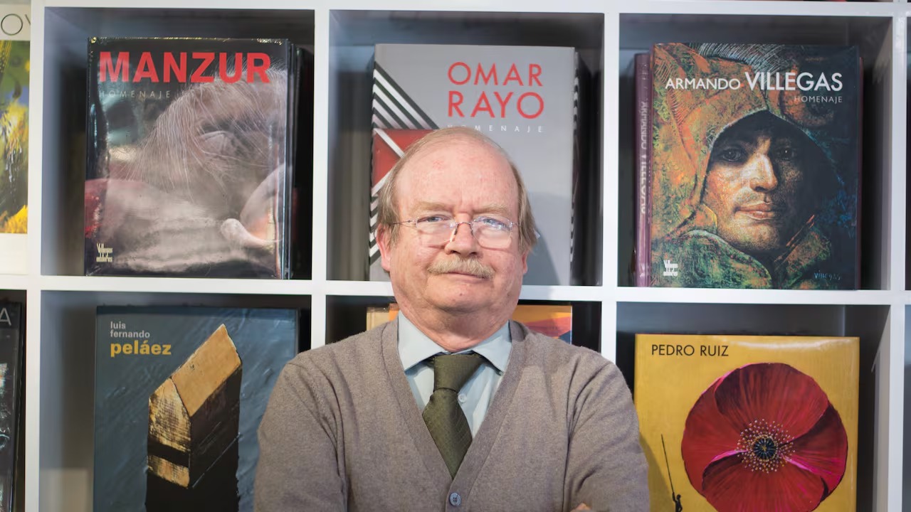Middle-aged man in a gray cardigan and tie stands in front of white shelves filled with large art books behind him. - Los lujosos libros de Villegas Editores que ahora podrán estar al alcance de cualquier bolsillo