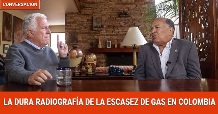 Two men sit across from each other at a wooden table in a cozy room, engaged in a conversation about gas scarcity in Colombia (banner text at bottom). - Colombia se queda sin gas y sin plan, la alerta que pocos quieren oír - Página 2