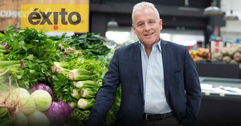 Man in a navy blazer and light blue shirt stands beside a fresh vegetable display in a market, with a yellow éxit sign behind him. - Carlos Mario Giraldo soportó el golpe laboral por la llegada de los Calleja, hasta alcanzar su jubilacion