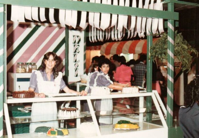 Two young women in blue uniforms stand behind a glass cake display at a retro bakery stall, with cakes arranged inside and striped decor above. - La repostería Deli le ganó a una multinacional China