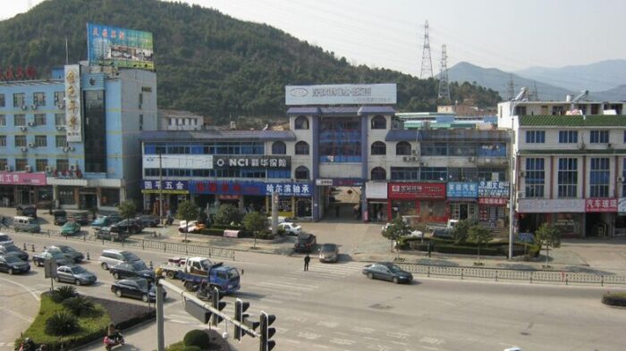 A busy street in a Chinese town with multi-story storefronts, parked cars, and hills in the background. - La repostería Deli le ganó a una multinacional China
