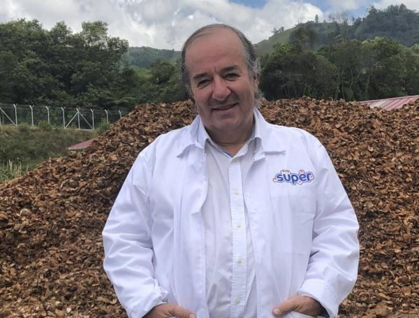 Man in a white lab coat standing outdoors in front of a large pile of wood chips, with hills and trees in the background. - La repostería Deli le ganó a una multinacional China