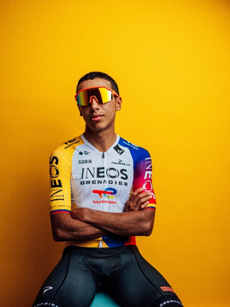 Male cyclist wearing Ineos Grenadiers jersey and orange goggles, arms crossed, against a solid yellow backdrop. - La jugada de Germán Bahamón para traer de vuelta Café de Colombia con equipo de ciclismo de talla mundial