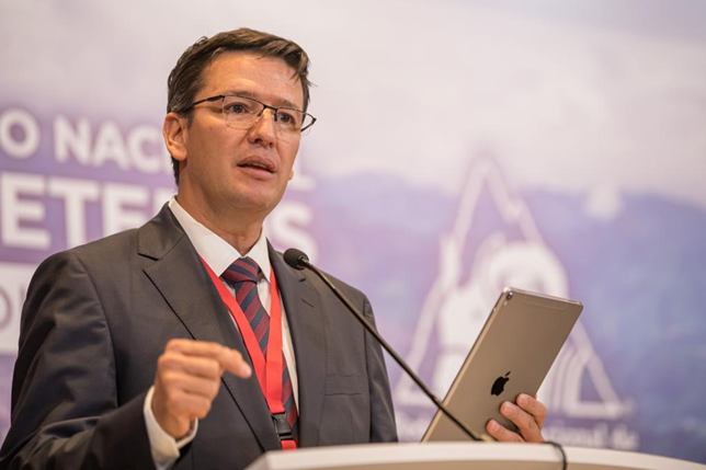 Businessman in a suit giving a conference presentation, gesturing with one hand while holding a tablet at a podium with a microphone nearby. - La jugada de Germán Bahamón para traer de vuelta Café de Colombia con equipo de ciclismo de talla mundial