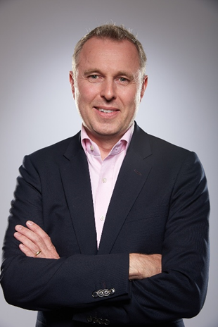 Head-and-shoulders portrait of a smiling man in a navy suit with a pink shirt, arms crossed. - La jugada de Germán Bahamón para traer de vuelta Café de Colombia con equipo de ciclismo de talla mundial