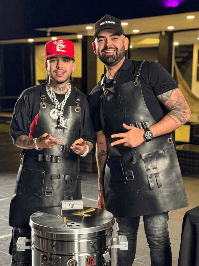 Two men in black leather aprons pose for a photo indoors; one wears a red cap and chunky chain, the other a black cap with visible tattoos. - La historia del paisa Andrés Humo Barriles y como logró exportar US millones con sus barriles para asar carne
