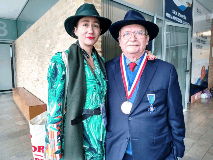 Woman in a teal-green patterned dress and hat stands with an older man in a navy suit and medal, posing in a modern building lobby. - Los lujosos libros de Villegas Editores que ahora podrán estar al alcance de cualquier bolsillo