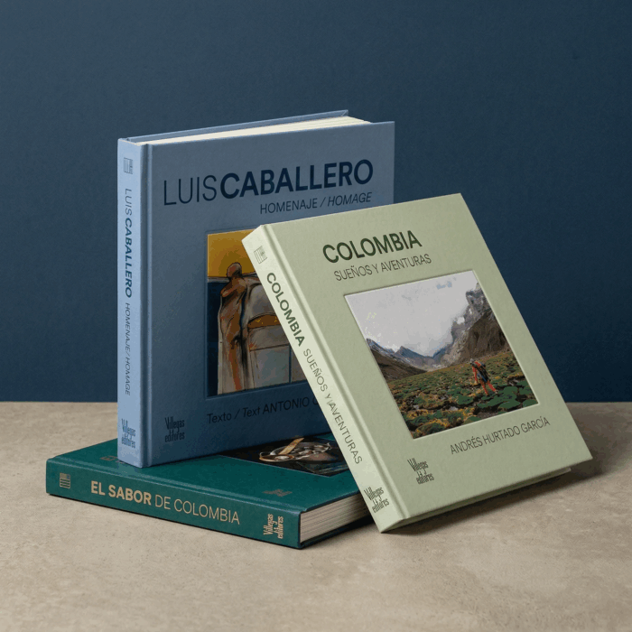 Stack of Spanish-language books about Colombia and travel, including 'El Sabor de Colombia' (teal), 'Colombia: Sueños y Aventuras' (light green), and blue 'Luis Caballero' volume. - Los lujosos libros de Villegas Editores que ahora podrán estar al alcance de cualquier bolsillo