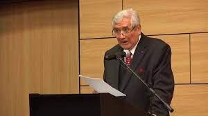 Older man in a dark suit and red tie speaks at a podium, reading a paper. - El lado costeño de Iván Cepeda por su abuela Aurita Chadid que lo hace familia de los Guerra, los Name y otros