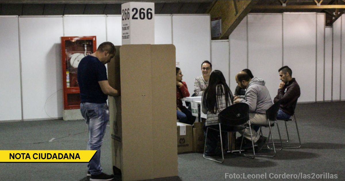 Man stands beside a tall cardboard voting booth while a group sits around a table with papers in a large hall; banner reads NOTA CIUDADANA. - El enorme descuadre fiscal al que tendrá que enfrentarse el próximo presidente de Colombia