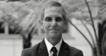 Portrait of an older man in a dark suit and tie wearing glasses, outdoors with palm fronds in the background, smiling softly. - Como creemos, así es que se puede