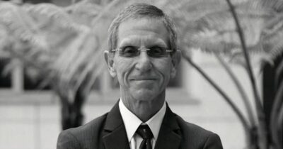 Portrait of an older man in a dark suit and tie wearing glasses, outdoors with palm fronds in the background, smiling softly. - Como creemos, así es que se puede