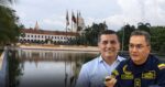 Two men stand in front of a colonial government building with a reflecting pool and flagpoles; one wears a light shirt, the other a navy uniform with patches and insignia, holding a small device; - La estratégica alianza entre la Armada Nacional y el alcalde Dumek Turbay para cambiar el Parque La Marina