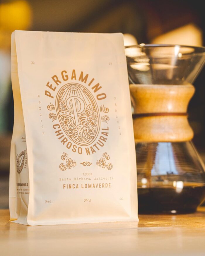 Bag of coffee beans with an ornate logo and text on a white bag, beside a glass pour-over brewer on a counter. - El Café Pergamino que pasó de conquistar Medellín a vender en 20 países, le apuesta a Canadá