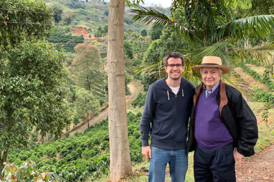 Two men posing outdoors in a terraced hillside farm, one in a hoodie and jeans, the other in a hat and purple sweater. - El Café Pergamino que pasó de conquistar Medellín a vender en 20 países, le apuesta a Canadá