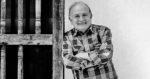 Older man in a plaid shirt leaning on a wooden railing, smiling at the camera (black and white). - Jorge Iván puede con la EPS