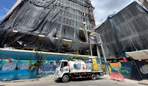 Construction site with a tall building wrapped in black tarps and scaffolding; a white service truck parked on the street in front. - ¿Quiénes están detrás de la Universidad Cooperativa, la institución que más gradúa abogados en Colombia?