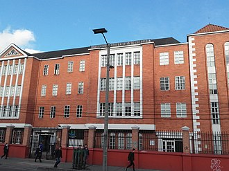 Red brick school building with white-framed windows and a central entrance; pedestrians walk along the sunny sidewalk in front. - ¿Quiénes están detrás de la Universidad Cooperativa, la institución que más gradúa abogados en Colombia?