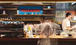 Busy cafe counter with baristas preparing drinks and customers ordering, warm lighting and shelves of cups in the background. - El Café Pergamino que pasó de conquistar Medellín a vender en 20 países, le apuesta a Canadá