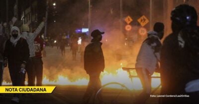 Nighttime street protest with masked demonstrators and flames burning in the road, overlaid with a yellow 'NOTA CIUDADANA' banner. - Las2orillas.co: Historias, voces y noticias de Colombia