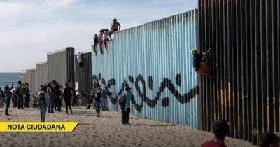 People climb and stand along a tall border wall on a sandy beach, with the sea to the left and bystanders nearby. - Las2orillas.co: Historias, voces y noticias de Colombia