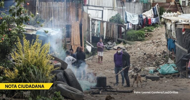 People stand in a dirt alley of a shantytown, smoke from a cooking fire rising, makeshift homes with laundry hanging above, debris on the ground. - El gran reto de De la Espriella es poder devolverle la tranquilidad a los territorios colonizados por la violencia