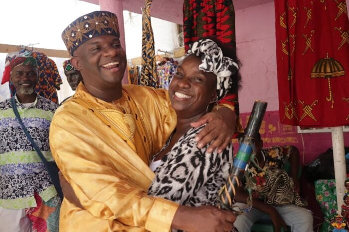 A man in a gold traditional outfit embraces a smiling woman wearing a zebra-pattern head wrap at a colorful market stall. - Un líder afro que avaló al papá de Miguel Uribe no quiso perderse la palomita de ser congresista por un mes