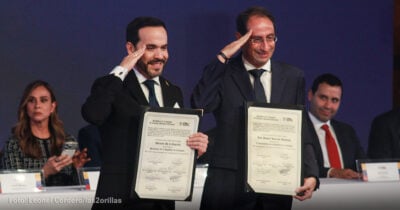 Two men in dark suits salute while holding framed certificates at a formal ceremony; a woman to the left watches on her phone. - Las2orillas.co: Historias, voces y noticias de Colombia