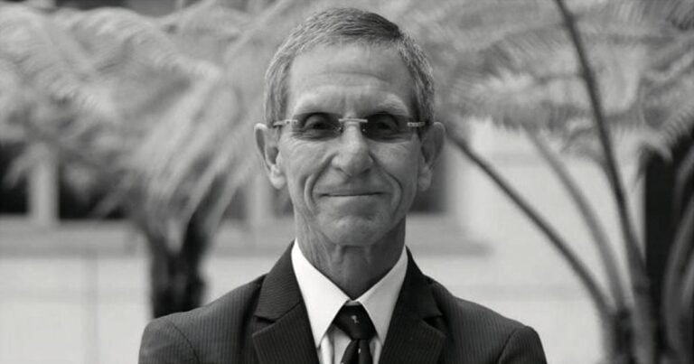 Older man in a dark pinstripe suit and tie, wearing glasses, smiling outdoors with blurred greenery in the background. - Los precios: entre la libertad y el control estatal