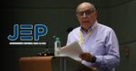 Older man in a light blue shirt stands at a podium reading from papers; a microphone and water bottle are on the lectern; left logo reads JEP. - El general de la guerra sucia en los Llanos aceptó responsabilidad ante la JEP