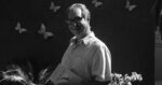Smiling man with glasses holding a book titled Medicina, with butterfly decorations in the background. - La salud va más allá de la biología