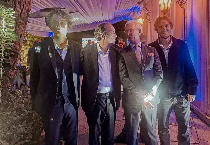 Cuatro hombres en trajes de negocios se encuentran juntos en un evento al aire libre, sonriendo y conversando bajo cálidas luces de cadena.