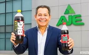 Smiling man in a suit holding two soda bottles, one labeled Kola Real, standing outside a building with an AJE sign in the background - Las poderosas familias peruanas que hacen fortuna en Colombia con gaseosas, colegios y cosméticos