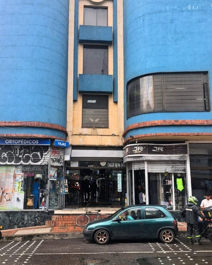 Así nació Distrito CH, la iniciativa que convirtió a Chapinero en un barrio amable Urban street scene with a bright blue curved building housing shops; a small green car parked in front and people on the sidewalk by storefronts labeled 'Ortopédicos' and fashion/storefronts nearby. - Así nació Distrito CH, la iniciativa que convirtió a Chapinero en un barrio amable