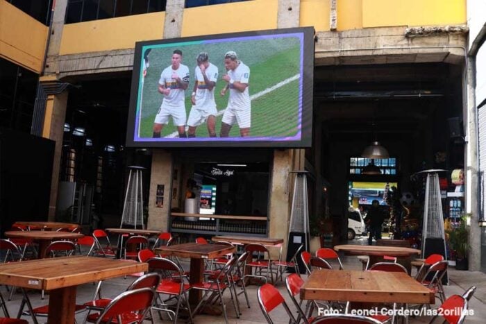 La antigua fábrica de Pastas El Gallo de donde salieron los primeros espaguetis a la venta en Bogotá sigue en pie Outdoor cafe with wooden tables and red chairs, facing a large mounted TV showing three soccer players in white uniforms. - La antigua fábrica de Pastas El Gallo de donde salieron los primeros espaguetis a la venta en Bogotá sigue en pie