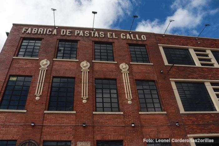 La antigua fábrica de Pastas El Gallo de donde salieron los primeros espaguetis a la venta en Bogotá sigue en pie Red brick pasta factory facade with tall windows and the sign 'FABRICA DE PASTAS EL GALLO' along the roofline on a sunny day - La antigua fábrica de Pastas El Gallo de donde salieron los primeros espaguetis a la venta en Bogotá sigue en pie