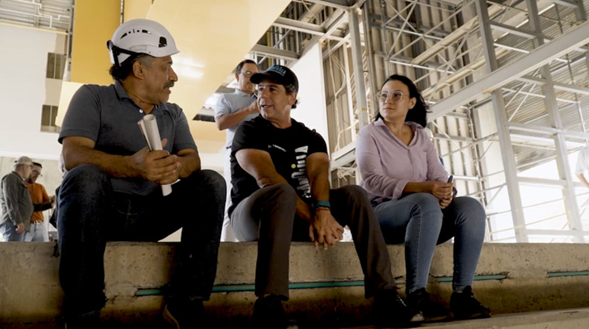 Three construction workers sit on a concrete ledge inside a building under construction, wearing hard hats and talking. - Tres constructoras que lograron levantar el Museo de Arte Moderno de Barranquilla