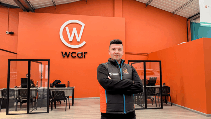 Man with arms crossed stands in front of an orange wall featuring the WCAR logo inside a modern office showroom. - Con la plataforma Wcar, dos hermanos bogotanos se metieron al negocio de los carros usados en Colombia