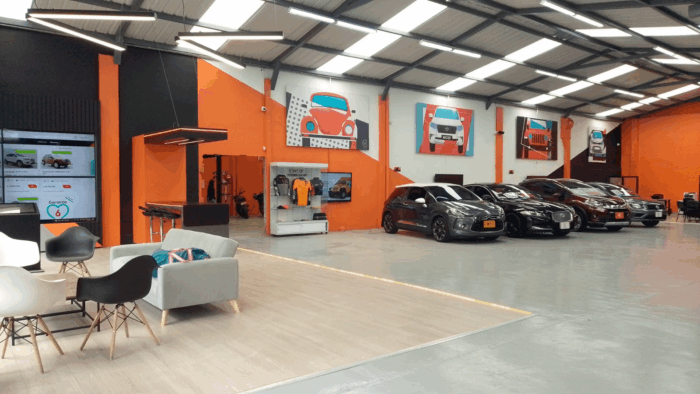 Interior of an orange-walled car showroom with several cars on display, a lounge seating area, and automotive artwork on the walls, left-side reception visible. - Con la plataforma Wcar, dos hermanos bogotanos se metieron al negocio de los carros usados en Colombia