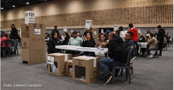 People seated at tables in a large hall voting area with ballot boxes and numbered signs above each station. - Así funcionará la plataforma que reportará irregularidades en tiempo real en las elecciones presidenciales
