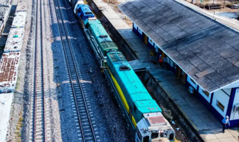 Overhead view of a turquoise-blue train at a small station, with a blue-roofed platform building on the right and a person waiting on the platform. - Con el sector privado detrás Ciénaga tendrá puerto moderno y moverá la carga del tren