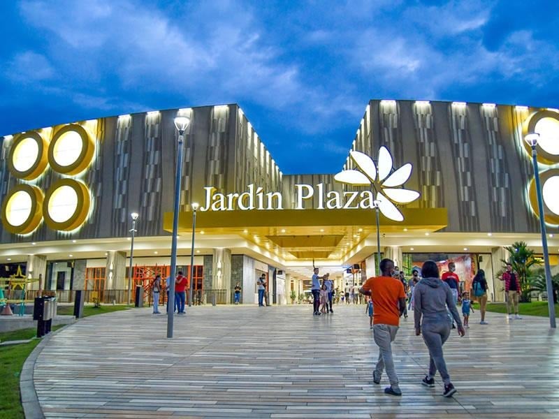 Exterior view of Jardín Plaza shopping center with people walking on a curved wooden walkway at dusk. - La apuesta de los chilenos de Cencosud para poner a competir su centro comercial en Cali contra los grandes