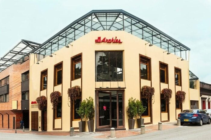 Después de 10 años, la cadena Archie’s vuelve a manos colombianas Two-story beige storefront with tall windows and a glass-roofed awning, sign reading 'Anchies' above the entrance, potted trees flanking the door, street scene with a blue car nearby. - Después de 10 años, la cadena Archie’s vuelve a manos colombianas
